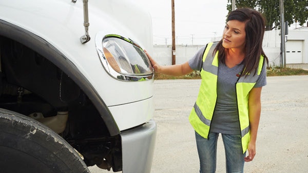 A commercial truck driver sits in the cab of a semi‑truck, hands on the steering wheel, approaching an intersection with traffic moving through multiple lanes on a clear day.