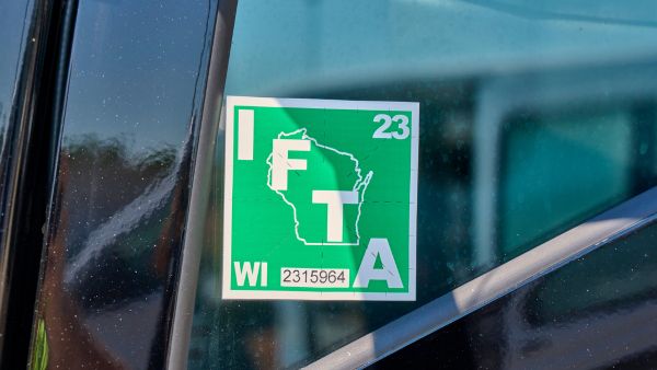 A green Wisconsin IFTA (International Fuel Tax Agreement) decal on a vehicle window, showing the outline of the state, the letters IFTA, and the WI identification number.